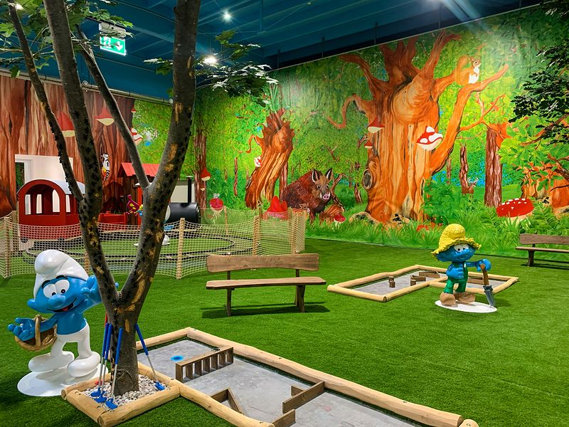 Minigolfbahnen auf grünem Kunstrasen, dazu Schlumpf-Figuren vor einem gemalten Wald. Foto: © Wolfgang DeMarco / ZIBINGU