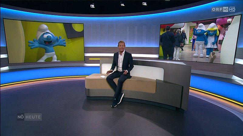 Standbild aus „Niederösterreich heute“: Moderator im ORF-Studio, auf den Bildschirmen Schlumpf-Figuren aus der Schlumpfwelt Traiskirchen